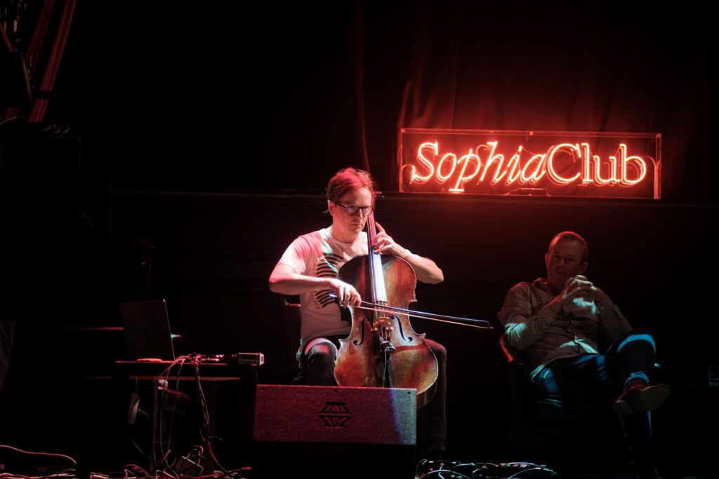 Photograph of cellist Oliver Coates performing onstage, with Tony Padilla watching behind and the Sophia Club sign in the background